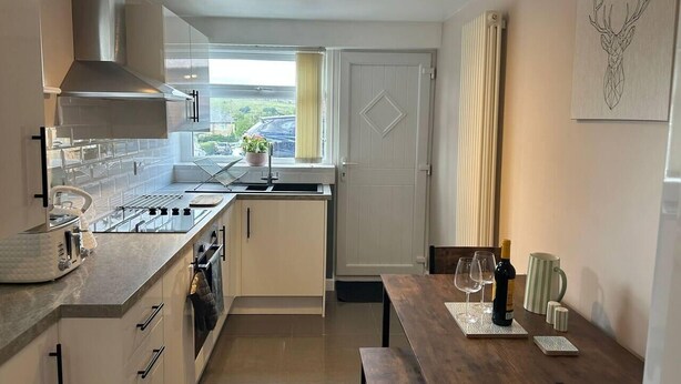 A compact kitchen with a gas stove, oven, and a modern range hood. There is a dining area with a wooden table and chairs, and a window with curtains. - 1-Bed House, Sleeps 4, Free Parking (Stockport)