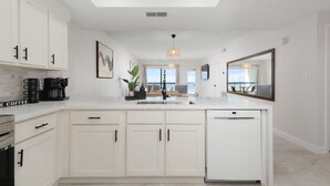 A modern kitchen with white cabinetry, a central island, a built-in dishwasher, and a large mirror above the sink. - Luxury beachfront condo with the best ocean view! (South Padre Island)
