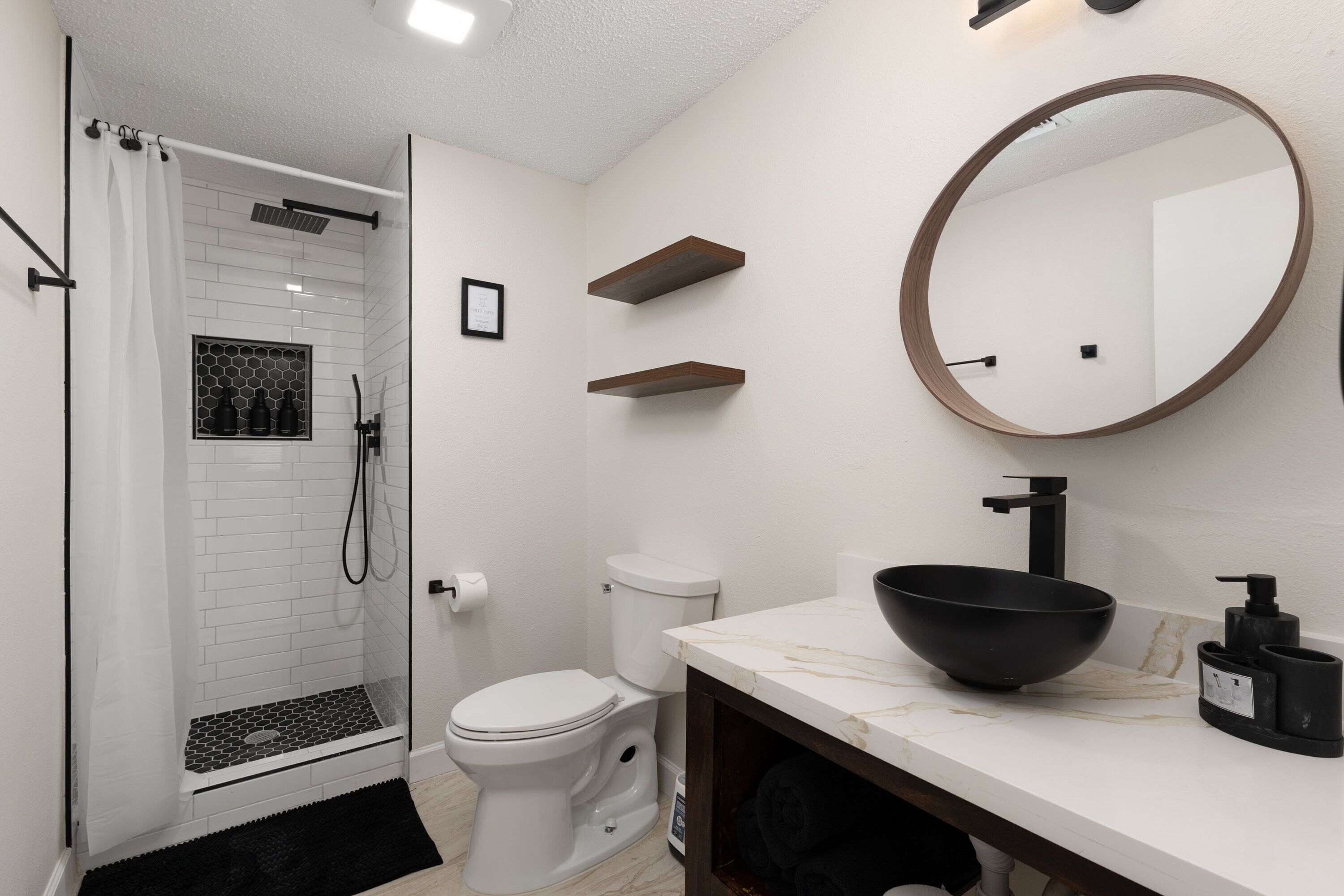 Et moderne badeværelse med en glasbrusekabine, et hvidt toilet, en marmorvask med en sort skål og et rundt spejl på væggen.