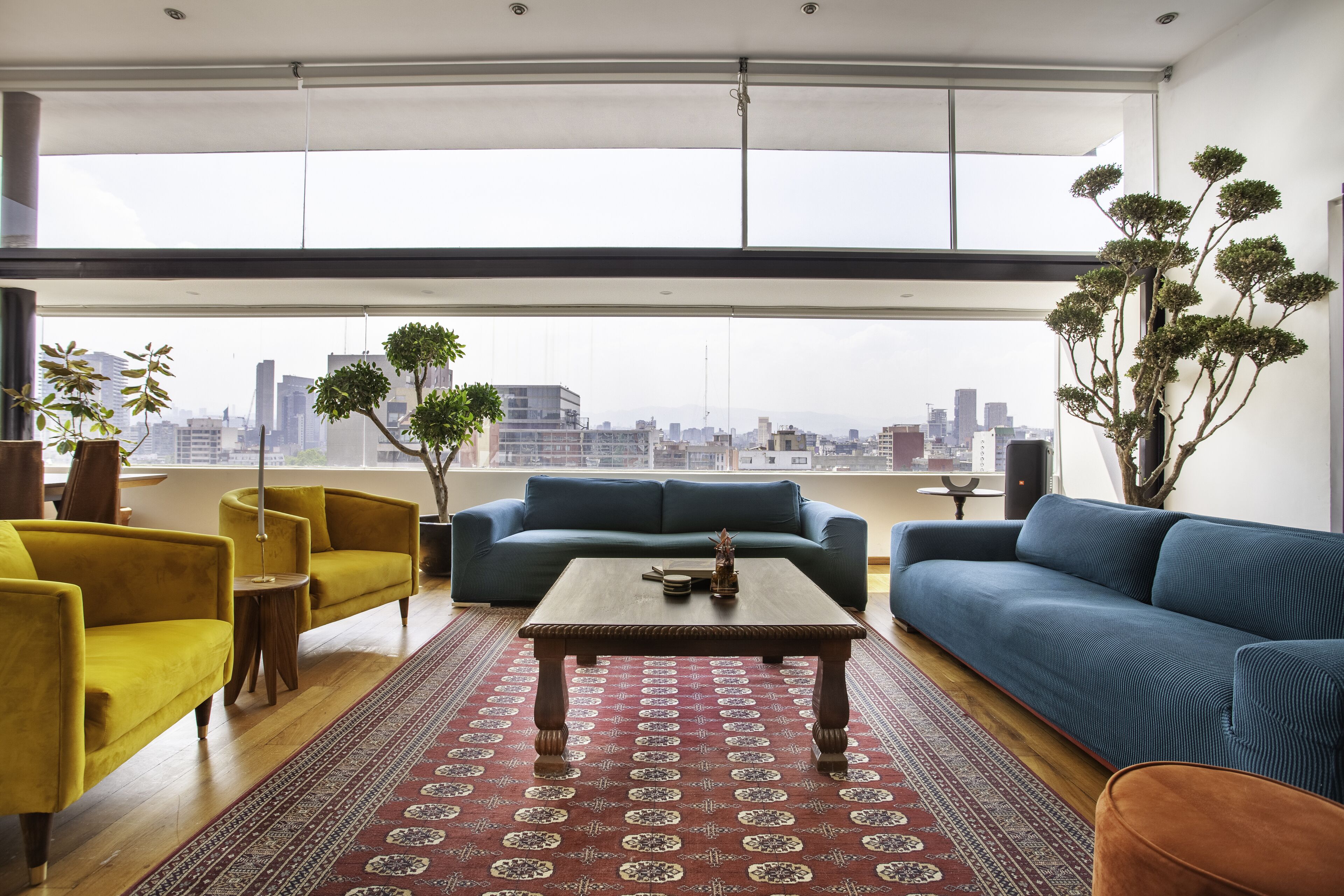 A modern living room with a city view, featuring a blue sofa, yellow armchairs, and a wooden coffee table.