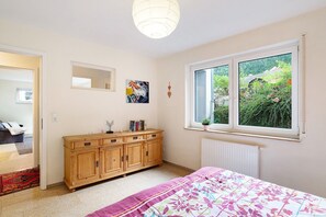 Une chambre à coucher avec un lit, une commode en bois, une fenêtre donnant sur un paysage verdoyant et un radiateur.