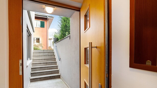 Ein schmaler Korridor mit einer Holztür, einer Treppe und Blick auf ein Gebäude mit einem grünen Fenster.