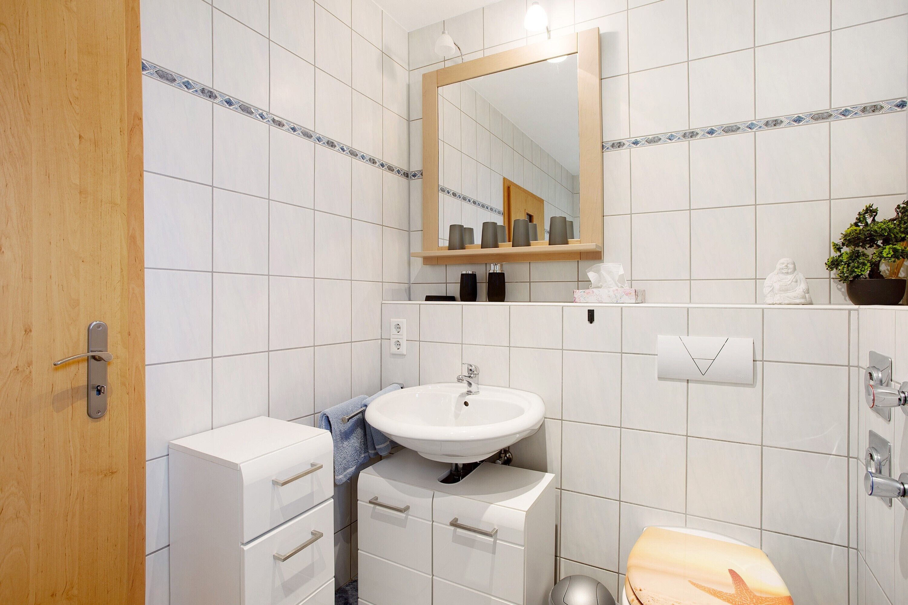 Un bagno con lavandino bianco, specchio e water. Si nota una porta in legno e un armadietto con cassetti.