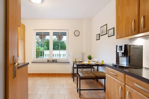 A kitchen with wooden cabinets, a dining table with chairs, a coffee machine, and a view through the window.