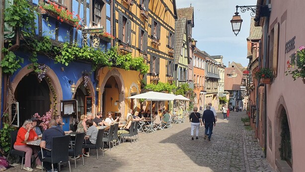 Een geplaveid straatje met terrasjes, traditionele vakwerkhuizen en een heldere lucht.