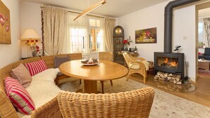 Een gezellige woonkamer met een ronde houten tafel, rieten meubels, een open haard en een klok aan de muur.