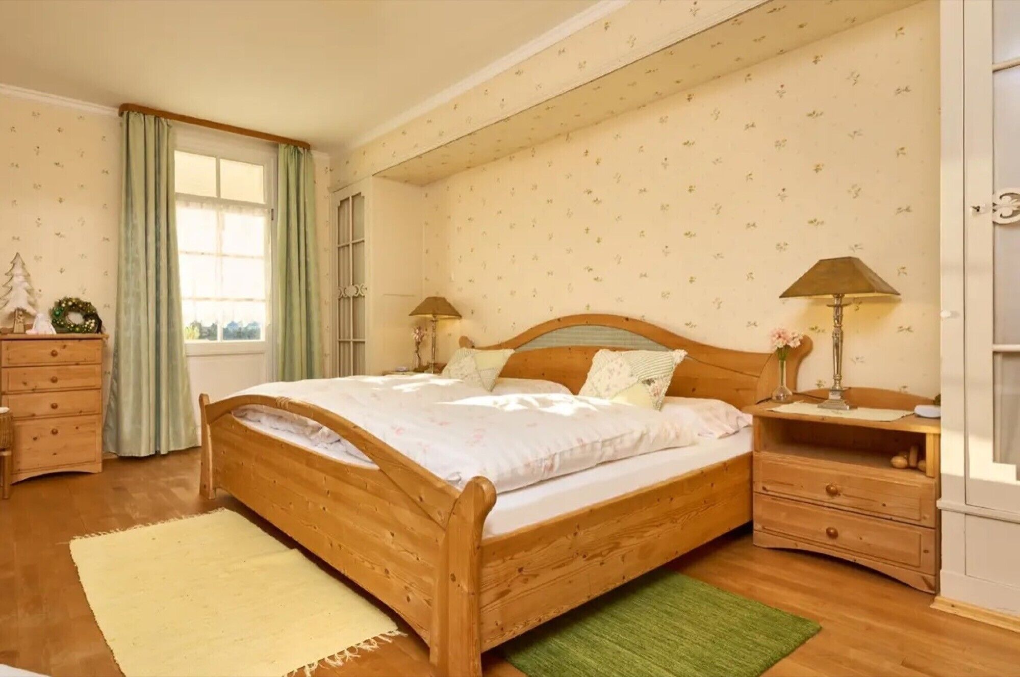 Una cama de madera con cabecera, dos mesitas de noche con lámparas, un aparador, una ventana con cortinas y una alfombra verde.