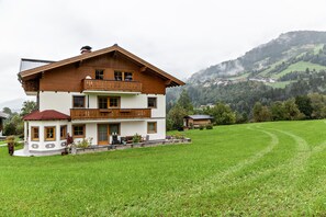 Ein zweistöckiges Haus mit Balkon, umgeben von einem grünen Feld und Hügeln im Hintergrund.