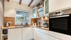 Une cuisine moderne avec des meubles blancs, un four encastré et une fenêtre donnant sur une maison et des arbres.