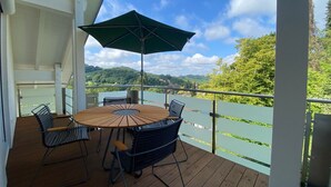 Une terrasse en bois avec une table ronde et des chaises, un parasol vert et une vue sur un paysage verdoyant avec des collines.