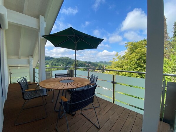 A wooden deck with a round table and chairs, a green umbrella, and a view of a green landscape with hills.