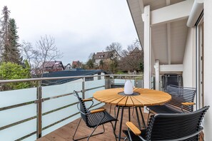 A balcony with a round wooden table and black metal chairs, overlooking a residential area with houses and trees.