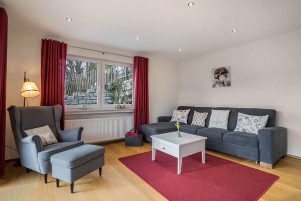 Een woonkamer met een grijze bank, een grijze fauteuil, een witte salontafel en een rood vloerkleed.