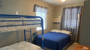 Una habitación con dos literas, una cama individual, un equipo de aire acondicionado, una ventana con cortinas, un ventilador de pie y una pequeña mesa de madera con un control remoto.