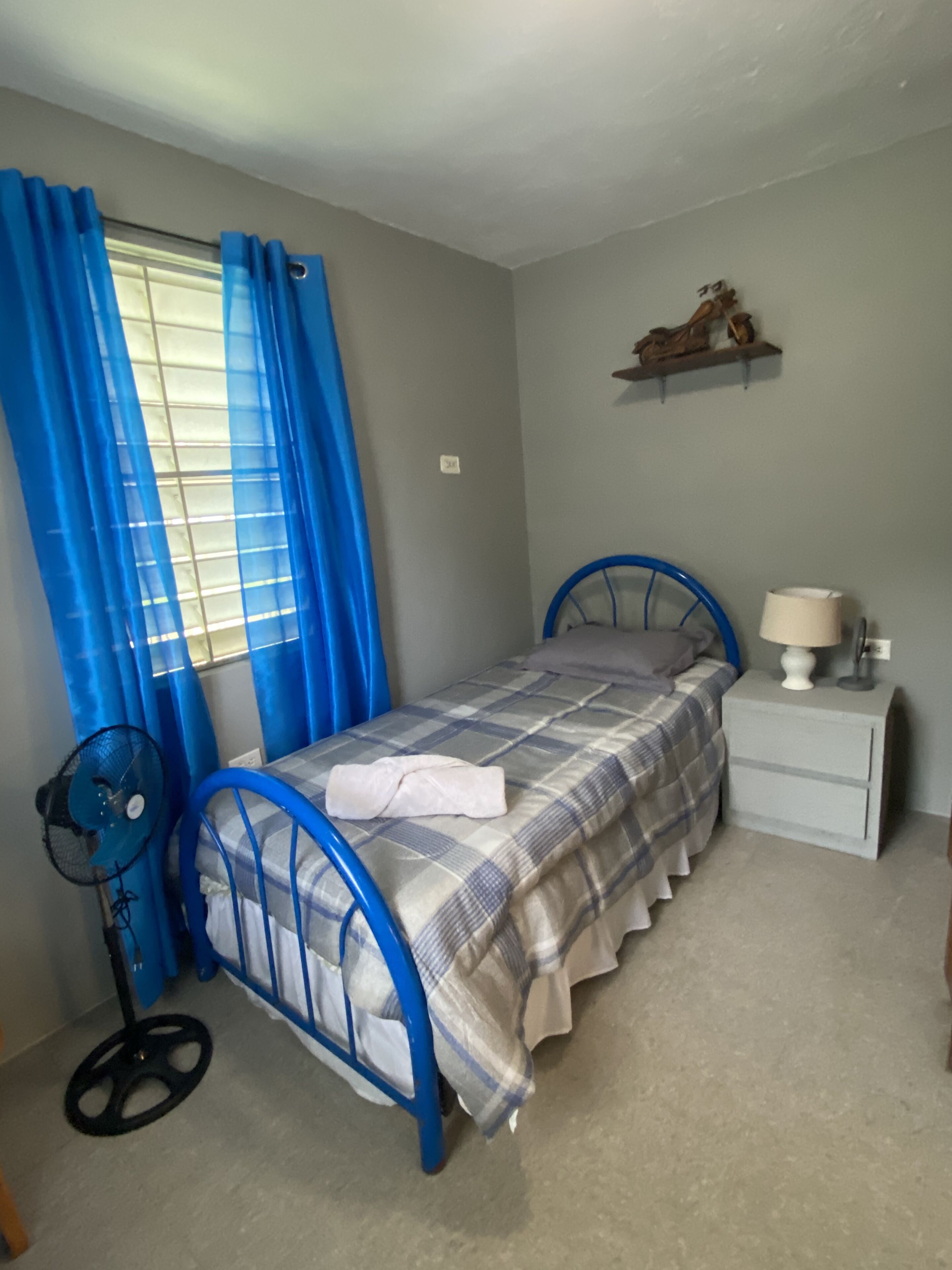 Un dormitorio con cama, mesita de noche, lámpara, ventilador y ventana con cortinas azules.