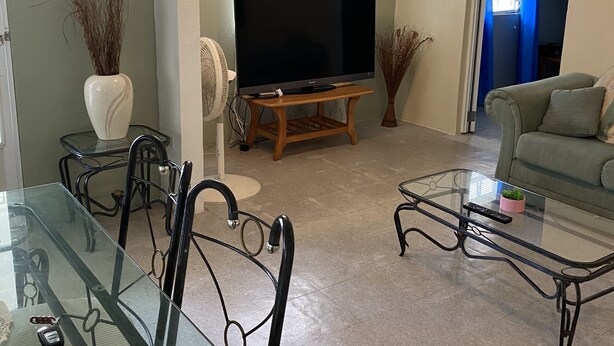 Una sala de estar con una mesa de vidrio para comer, sillas, un sofá, un televisor sobre un mueble bajo y una puerta que da a otra habitación.
