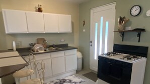 Cocina compacta con armarios blancos, estufa a gas, lavaplatos y una zona de comedor con una silla.