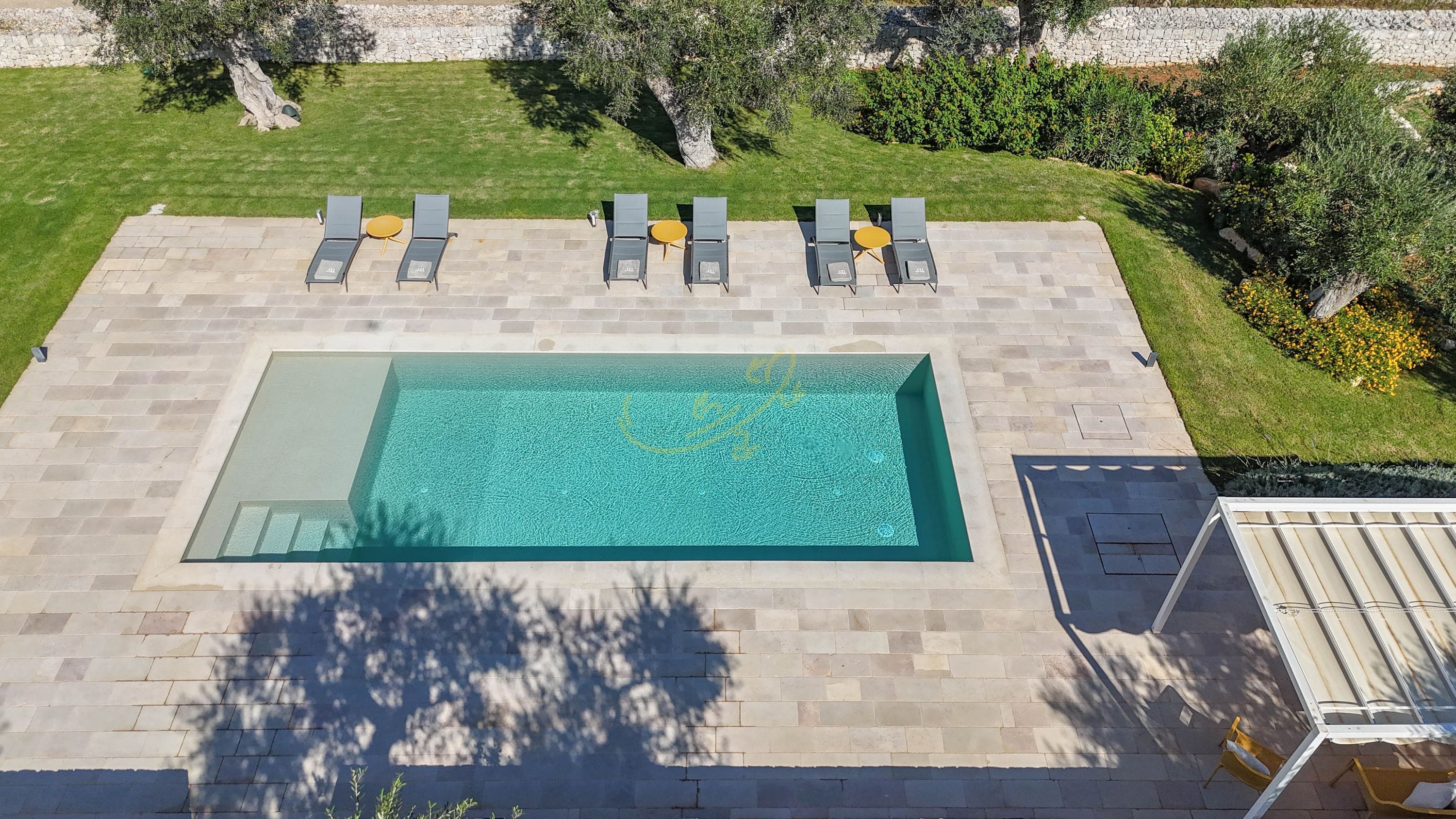 An outdoor swimming pool with a tiled deck, surrounded by lounge chairs and a stone wall.