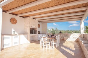 An outdoor patio with a stone wall, a brick oven, and a dining area with white chairs and a table. - TD Trulli Lorusso Design Luxury Trulli With Pool (Castellana Grotte)