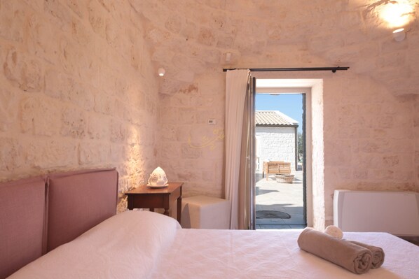 A bedroom with a stone wall, a bed with a headboard, a bedside table with a lamp, and a view of the outside. - TD Trulli Lorusso Design Luxury Trulli With Pool (Castellana Grotte)