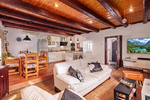 Un amplio espacio de sala con un sofá de cuero blanco, una mesa de comedor de madera y sillas. Se observa una cocina con gabinetes blancos y un ventanal grande al fondo.