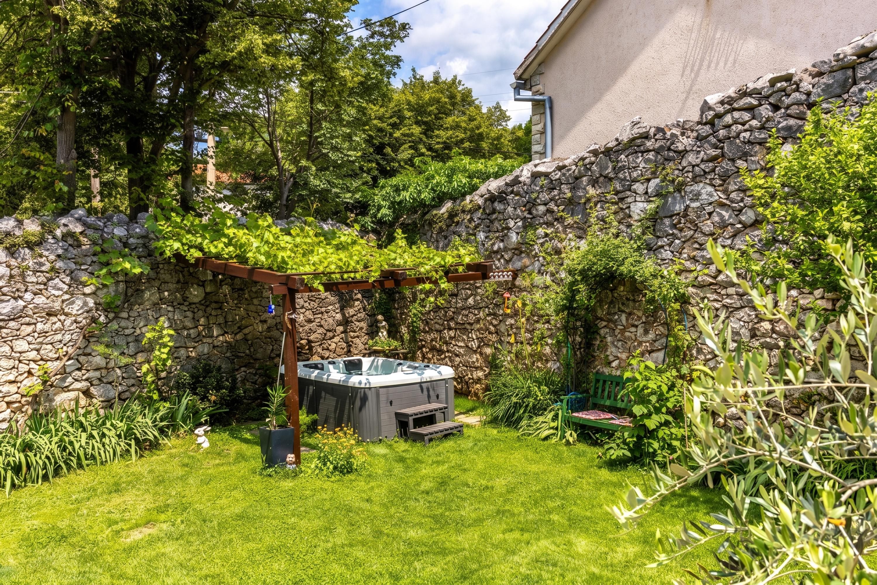 A backyard with a stone wall, a hot tub, and a pergola covered in greenery.