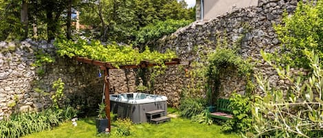 A backyard with a stone wall, a hot tub, and a pergola covered in greenery.