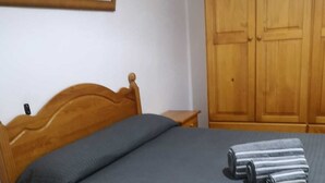 A bedroom with a wooden bed frame, a grey bedspread, a wooden nightstand with drawers, and a wooden wardrobe.