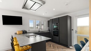 A modern kitchen with a central island, built-in appliances, and a large window. - The Paddock - Lovely Cottage With Hot Tub (Tenby)