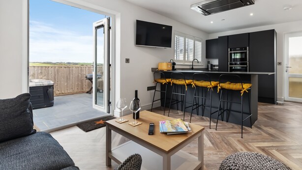 A modern kitchen with a flat-screen TV, a dining area with bar stools, and a large sliding glass door leading to an outdoor patio. - The Paddock - Lovely Cottage With Hot Tub (Tenby)