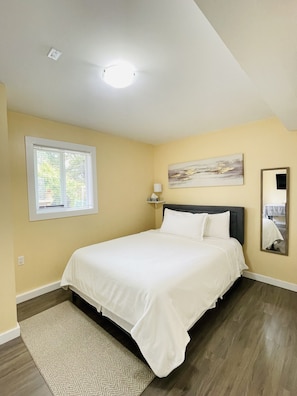 A bedroom with a bed, a window, a painting, a mirror, and a ceiling light.