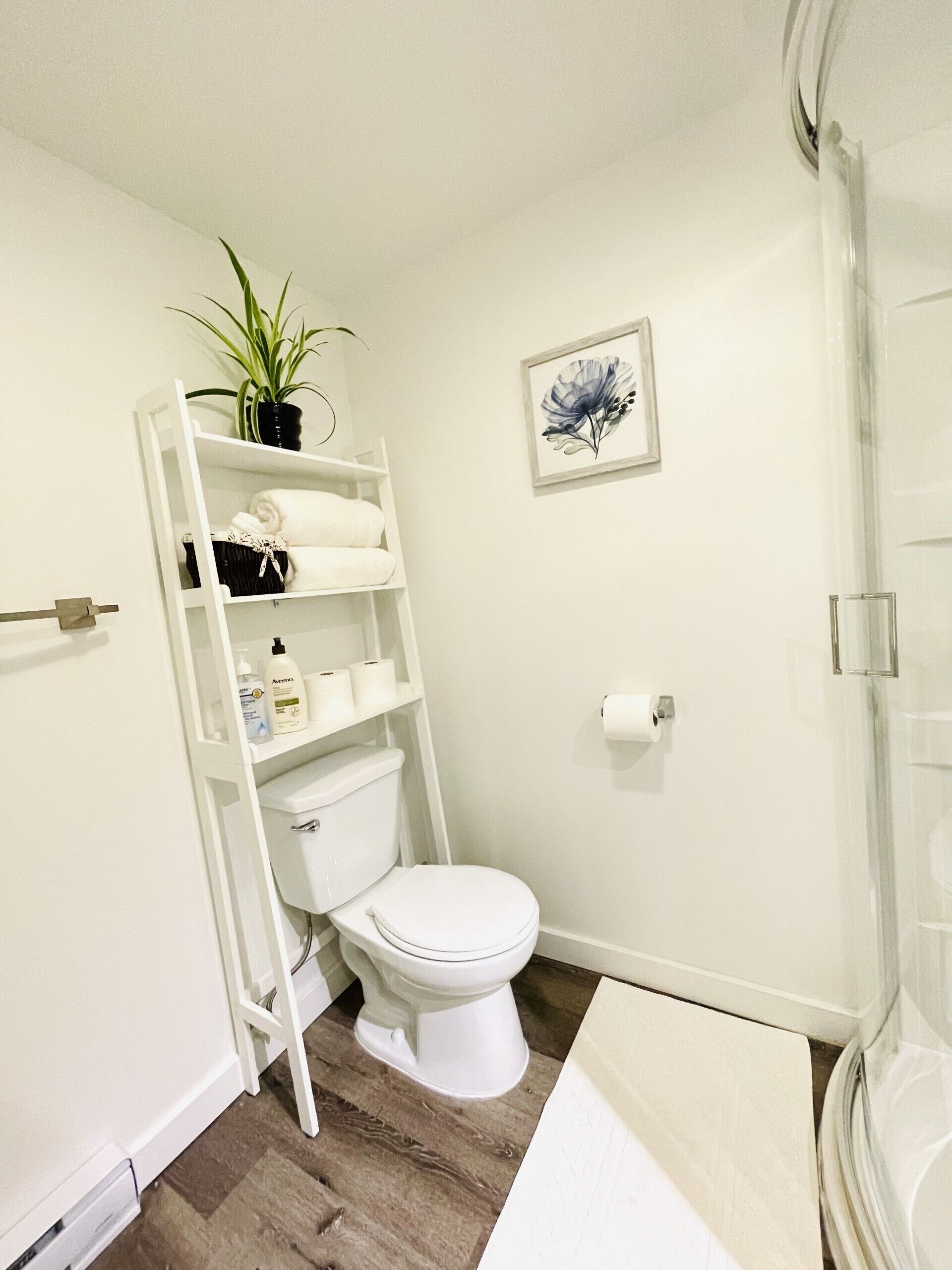 Une salle de bain blanche avec des toilettes, des étagères, une plante et un tableau encadré au mur.