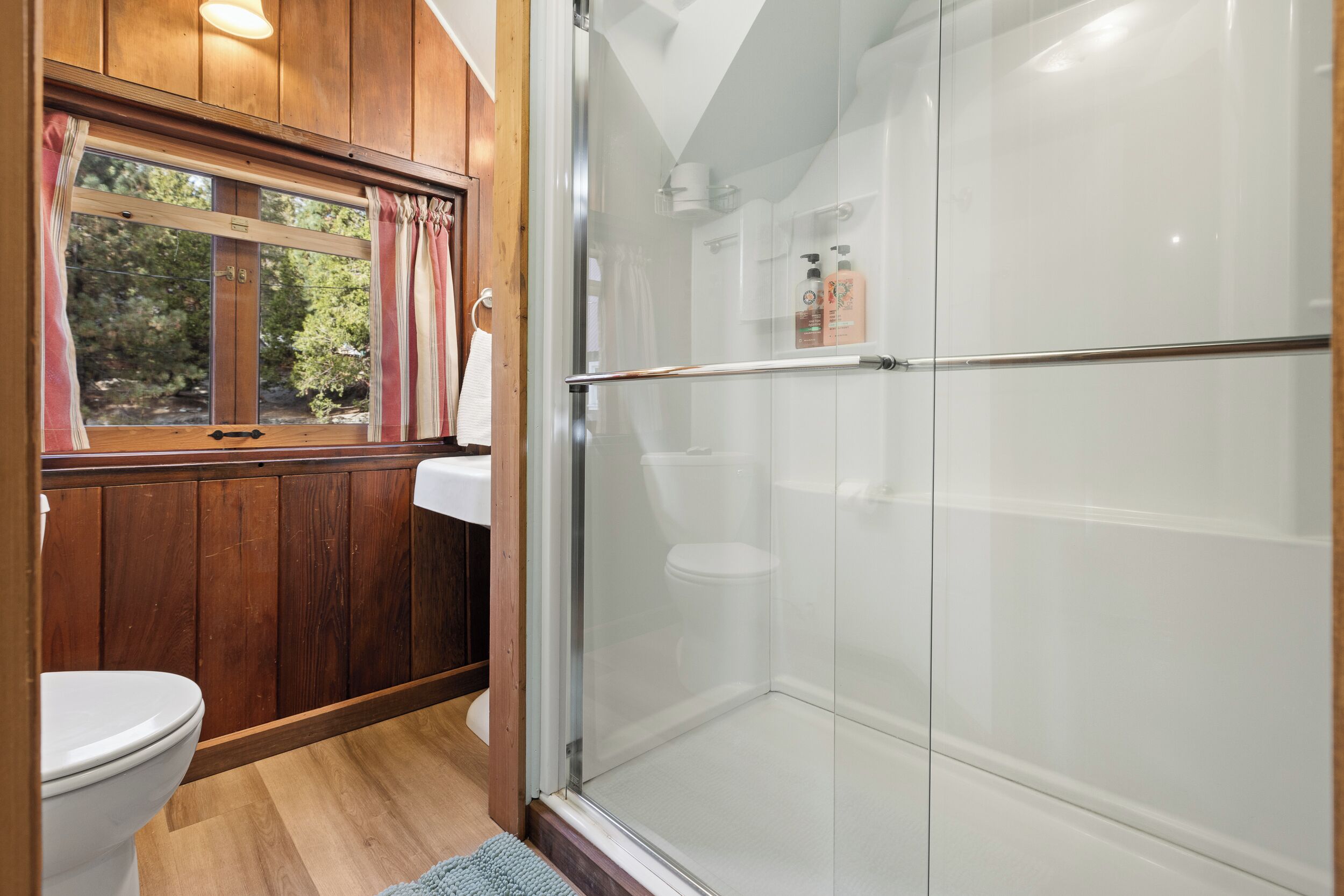 A bathroom with a wooden vanity, a toilet, a glass shower enclosure, and a window with curtains.
