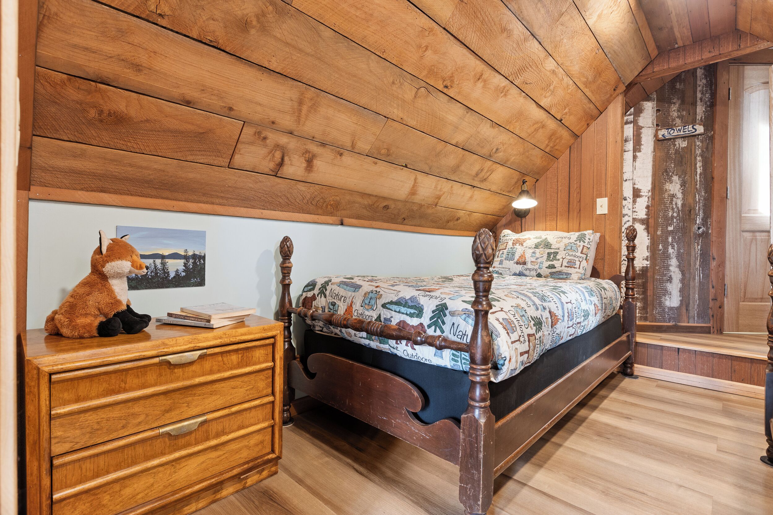 A wooden-paneled bedroom with a bed, a dresser, a lamp, and a stuffed toy.