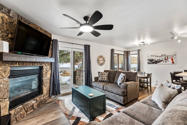 Cozy and modern living room with a rustic stone fireplace, wall-mounted TV, plush seating, and large windows offering a scenic, snowy view — perfect f