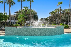 A swimming pool with a fountain, surrounded by palm trees and a tiled wall.