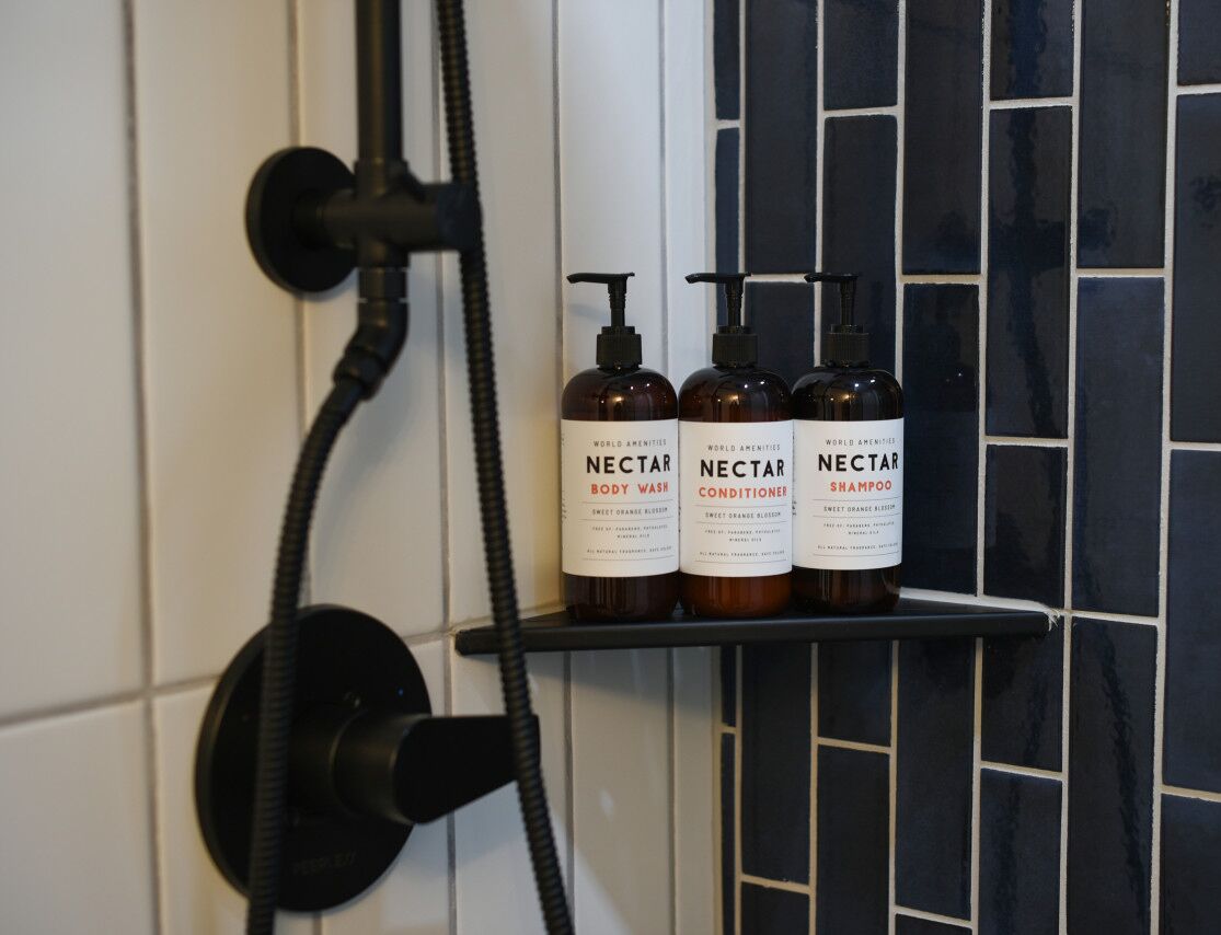 Een badkamer met een douche, een handdroger en drie flessen Nectar body wash, conditioner en shampoo op een plank.