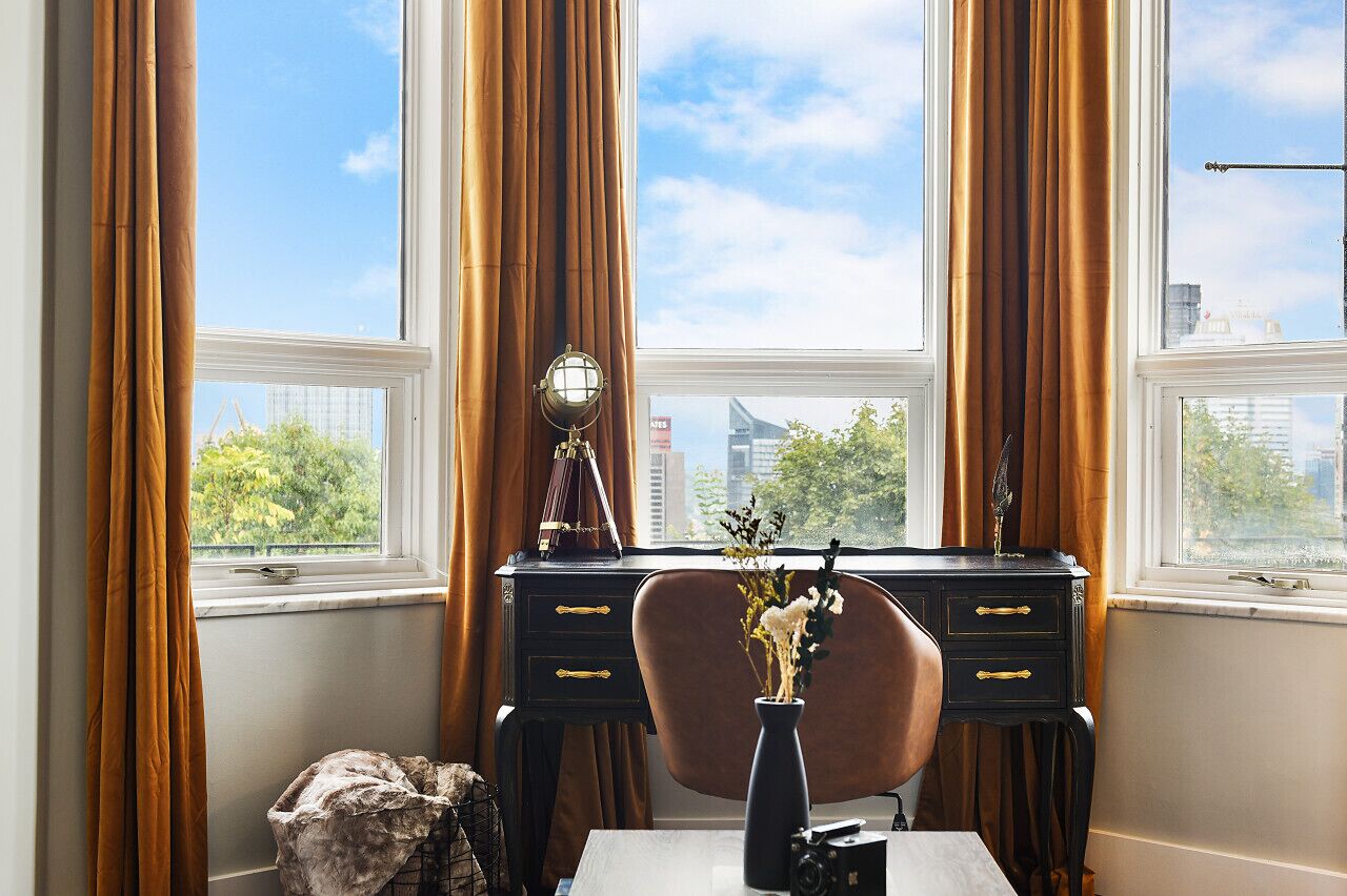 A room with a desk, chair, and a vase with flowers. There are two windows with curtains, and a cityscape is visible outside.