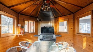 Interior de una cabaña de madera con una mesa central y sillas, un ventilador de techo y una zona de barbacoa.