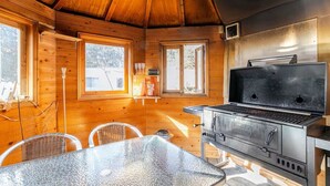 Interior de una cabaña de madera con una estación de parrilla metálica, una mesa de vidrio, sillas de mimbre y ventanas grandes.