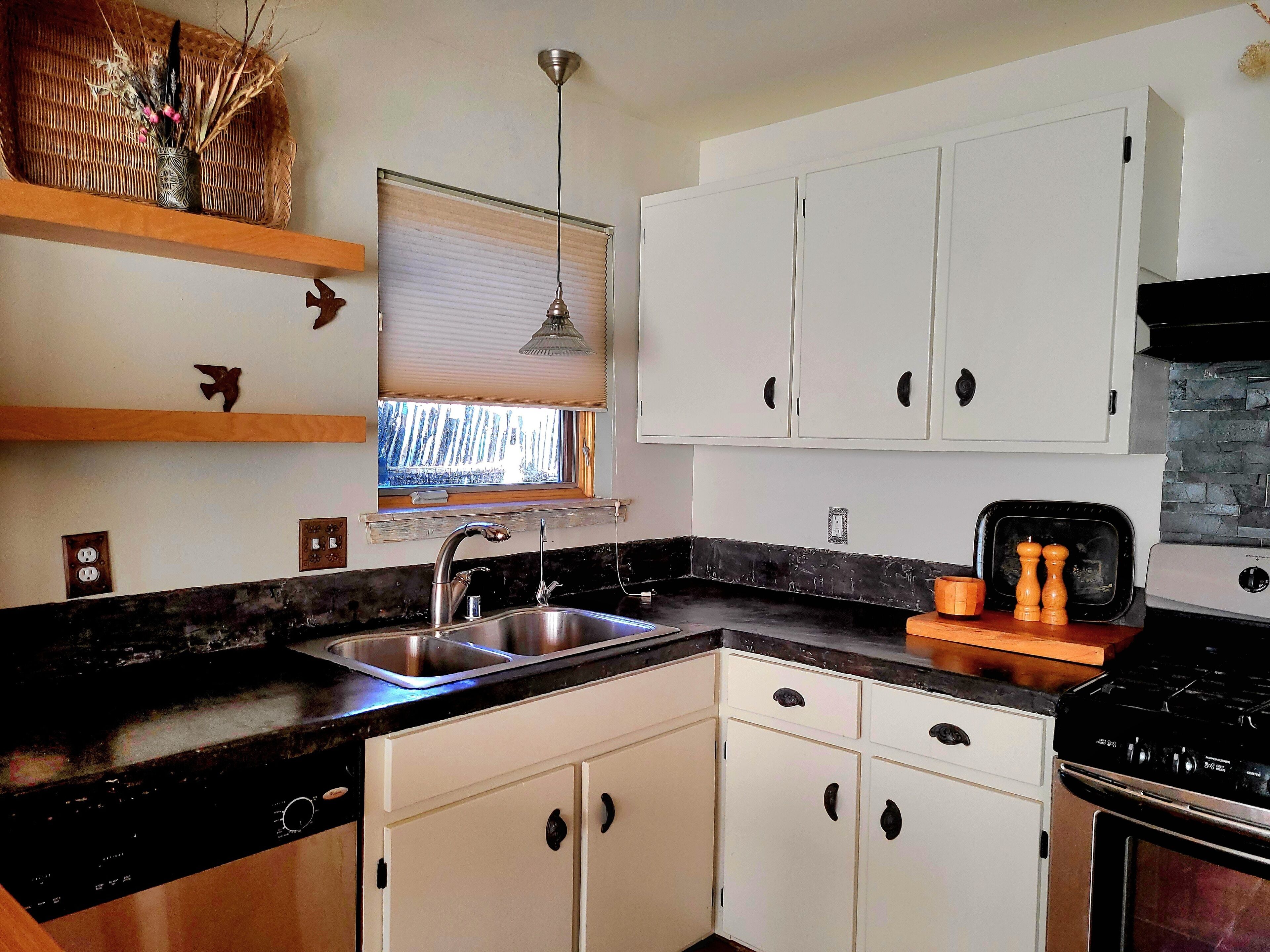 A modern kitchen with white cabinets, a black countertop, a stainless steel sink, a window with blinds, and a hanging pendant light.