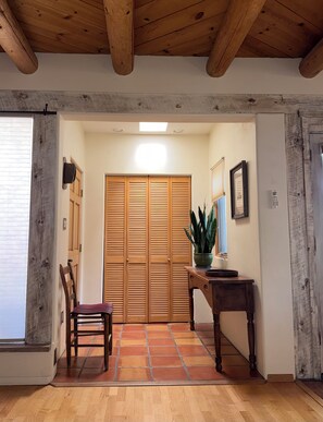 Un corridoio con soffitto in legno, pavimento piastrellato, una porta in legno, una sedia, un tavolo con una pianta in vaso e un punto luce a parete.