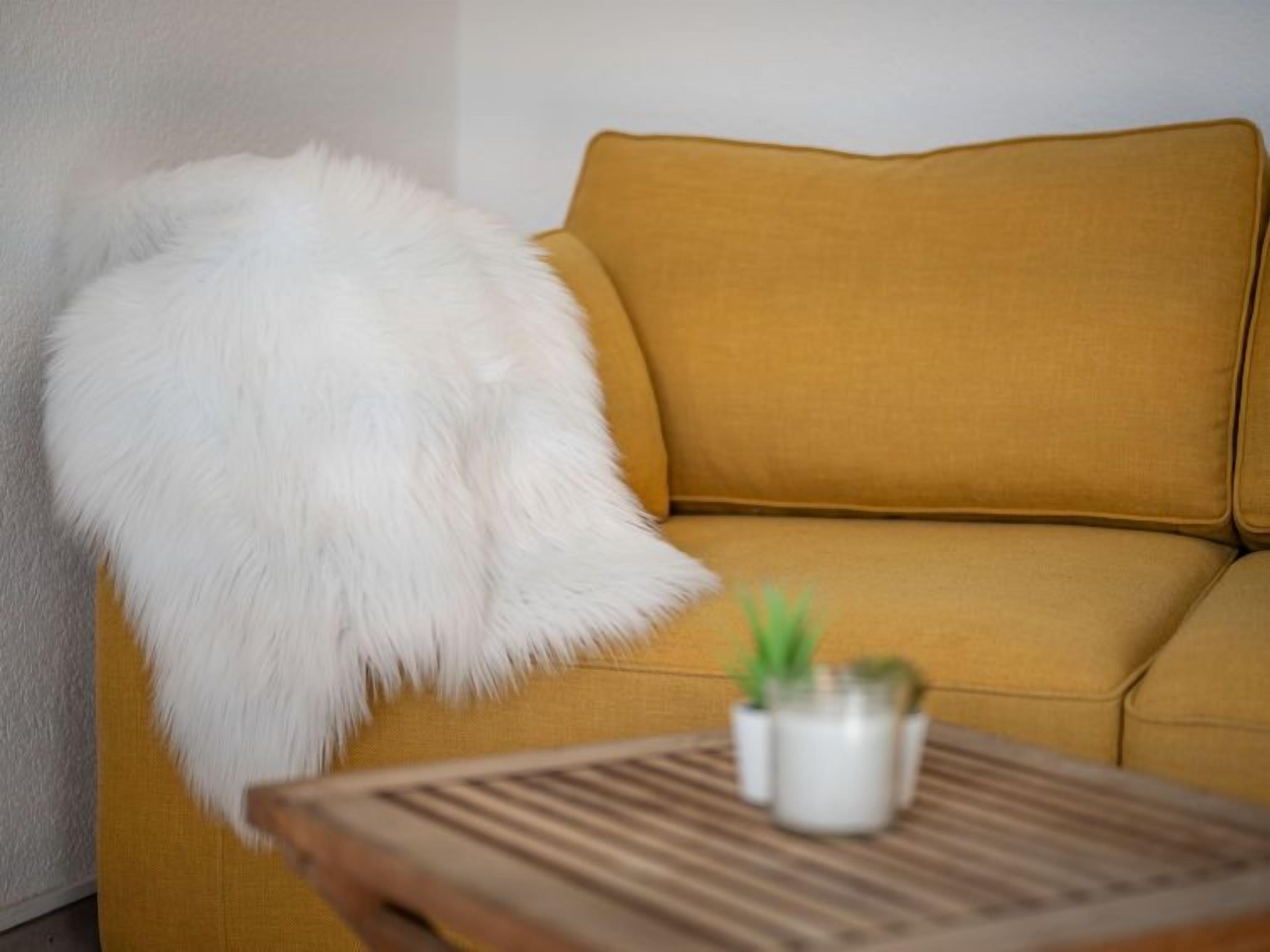 A mustard-colored sofa with a white fluffy throw, a small potted plant, and a wooden table with a striped pattern.