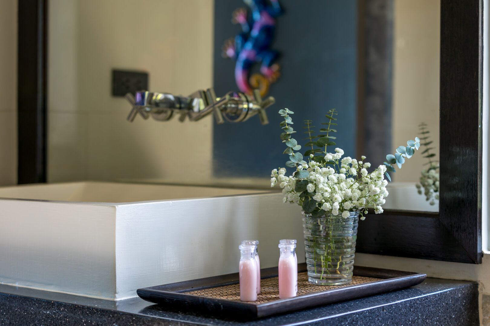 A bathroom with a bathtub, a mirror, and a tray with two bottles and a vase of flowers.
