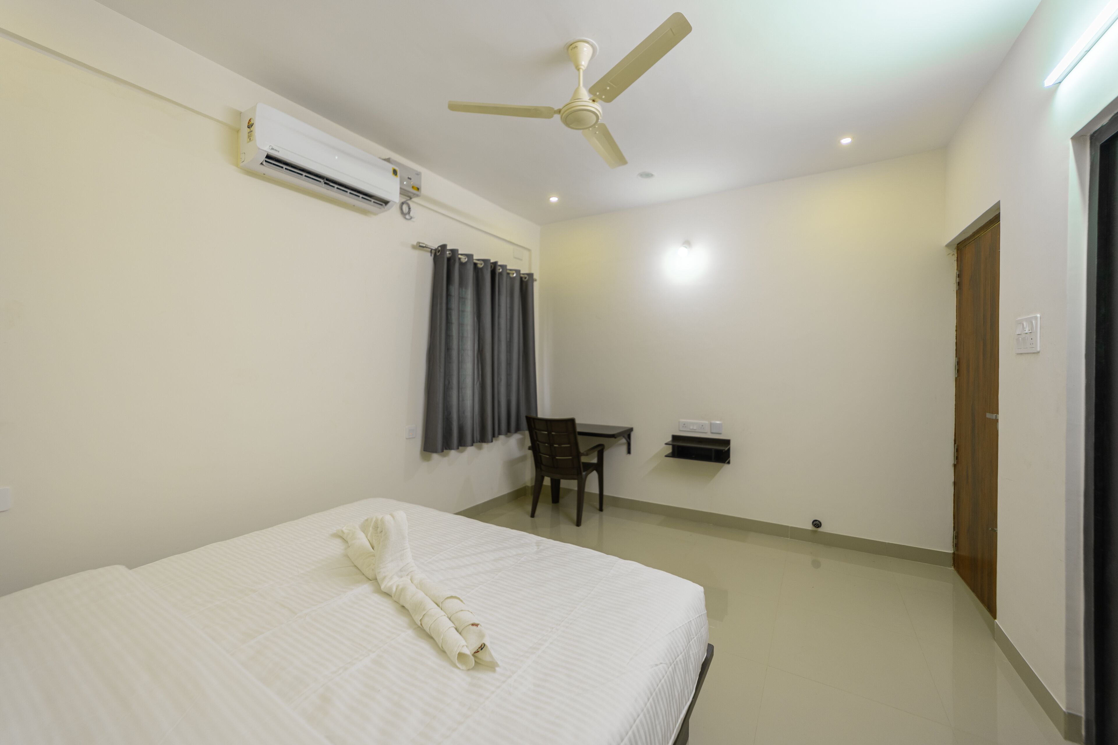 Una habitación de hotel con cama, escritorio, silla, ventilador de techo y aire acondicionado, además de una puerta.