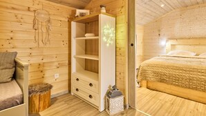 Um quarto de madeira acolhedor, com uma cama, um guarda-roupa branco, um pequeno banquinho e um candeeiro decorativo.