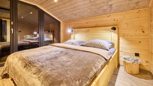 A wooden cabin bedroom with a large bed, a small wooden stool, and a glass door leading to another room.