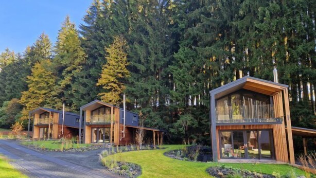 A modern cabin-style building with a glass balcony, surrounded by a forested area.