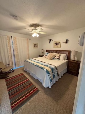 Un dormitorio con cama, mesita de noche, silla, ventilador de techo y ventana con cortinas.