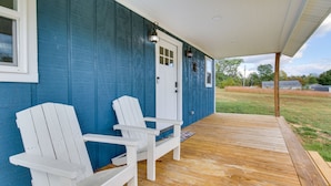 A porch with two white Adirondack chairs, a wooden deck, and a blue house with a white door.